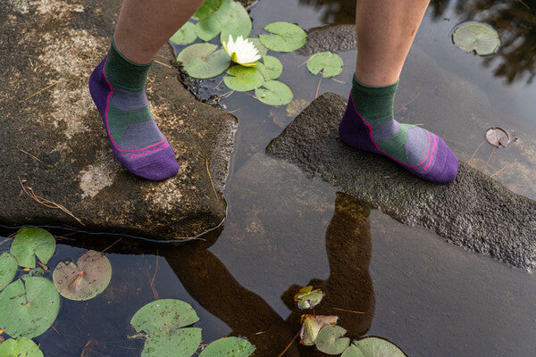 Feet crossing stream on rocks wearing moisture wicking merino wool socks in green and purple