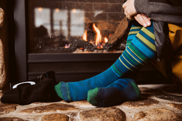 A person wearing blue darn tough socks, sitting next to a fireplace, looking cozy and comfy