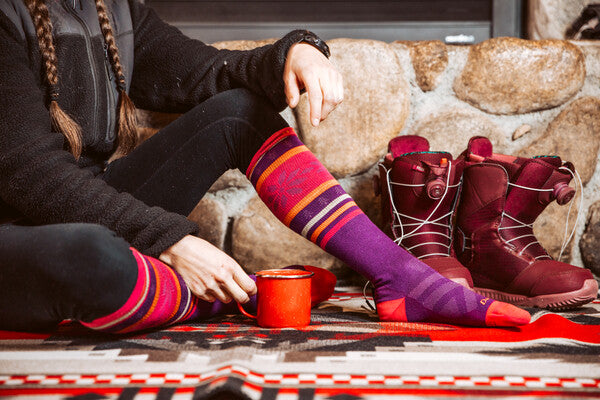 Person seated by fire wearing knee high darn tough socks in vibrant pink and purple
