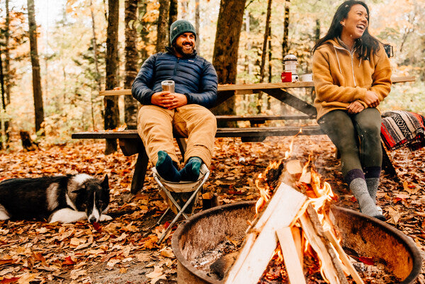 Two campers wearing twisted yarn boot socks seated by a campfire with their dog