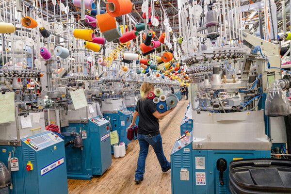 Darn Tough employee with arms full of yarn cones walking down between knitting machines