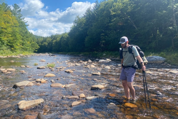 A hiker wading through a stream out in the mountains, getting his feet soaked