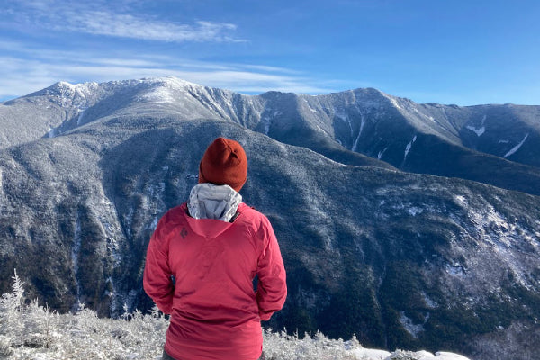 Hiker standing on summit of mountain wearing hat, coat, and surrounded by snow