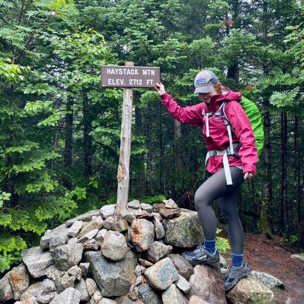 Alex wearing a raincoat standing at a summit ringed in by trees with the hiking sign