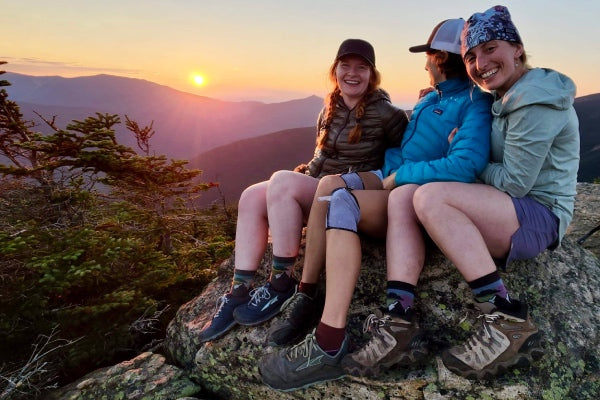Three hikers watching the sunset from a summit while wearing hiking socks with a sunset design
