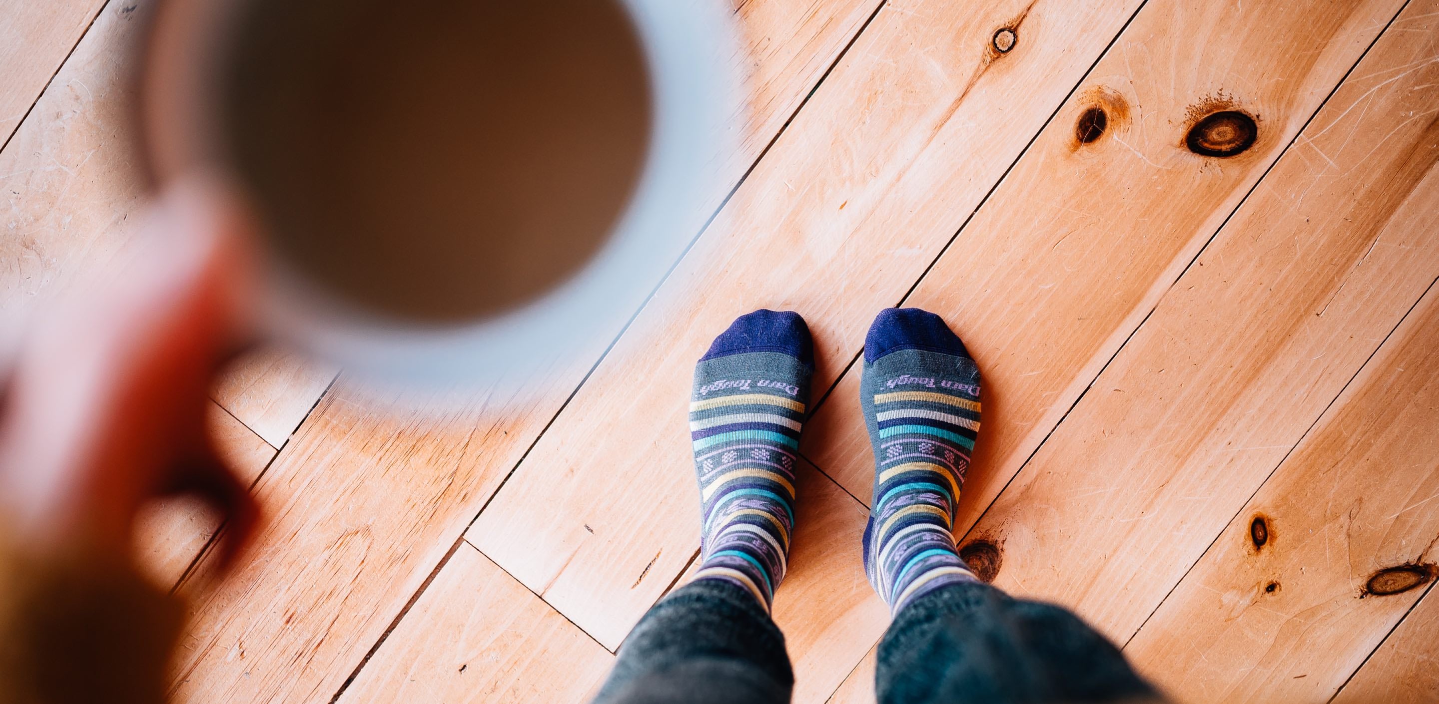 Person wearing Bronwyn lifestyle socks on a wood floor holding a cup of coffee