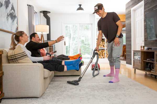 Two people seated on couch with sock-clad feet in the air while another person vacuums