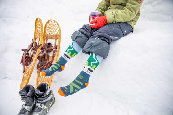 Model sitting with the Kid's Pow Cow Over-the Calf Snow Socks on a snow bank with hot chocolate next to snow shoes and boots