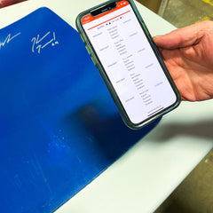Image of hand holding an iphone and showing the content on screen. Screen shows a webpage with a listing for a product with information about authenticated signatures and their dates. 