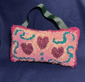 A small (6' x 9") pillow with a dark green grosgrain ribbon along the top-edge. The pillow has three pink hearts and some teal-colored swirls in the design.