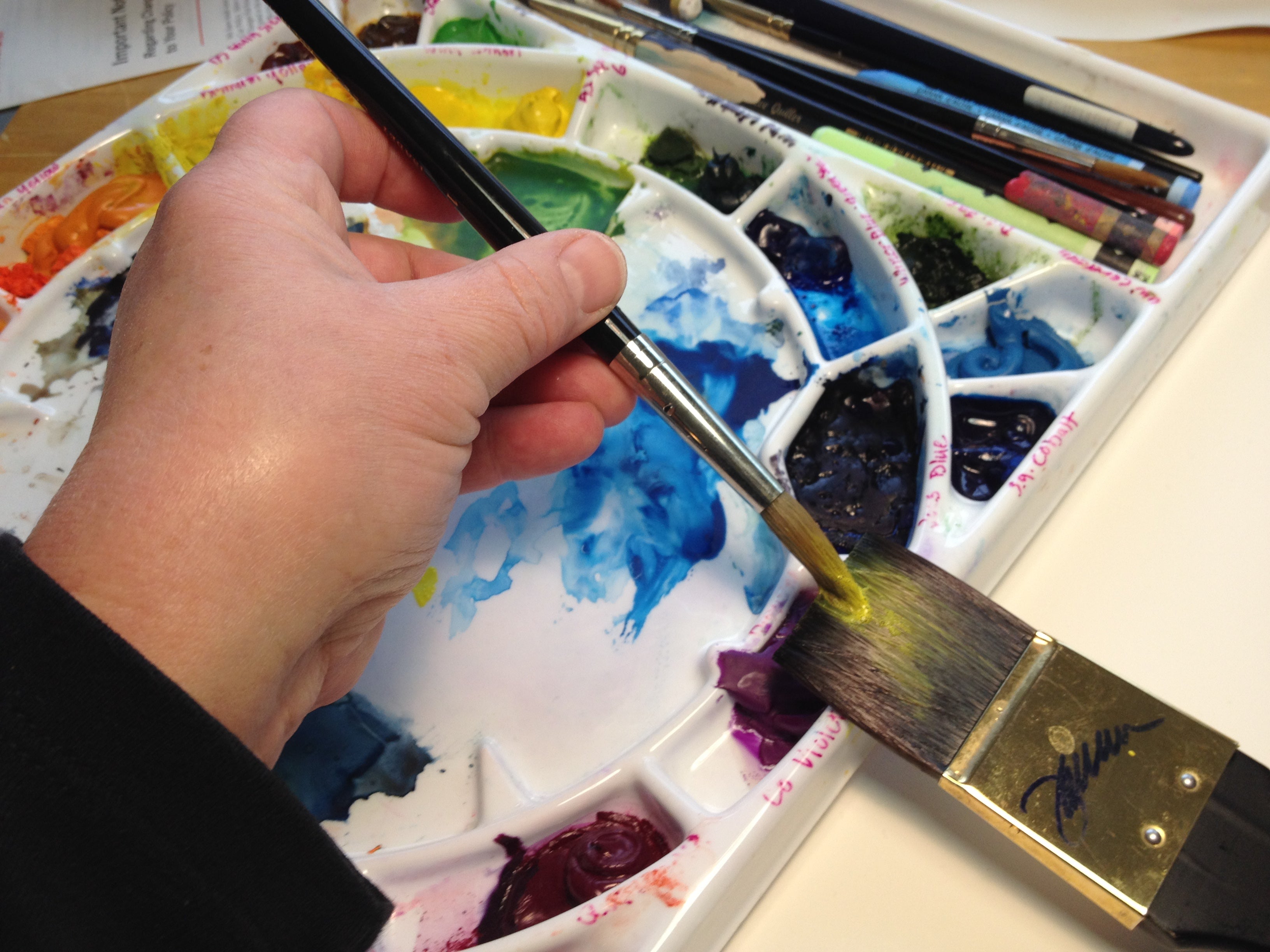 A photo of the left hand of the artist holding a brush as she applies paint to the bristles of a flat brush.