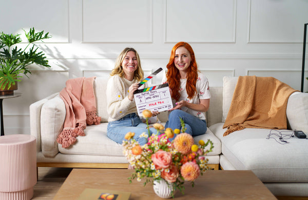 Natalie and Meg holding a clapper seated on a couch near flowers in a vase