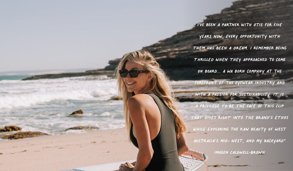 Imogen Caldwell Brown holding her surfboard on the beach and smiling wearing black sunglasses