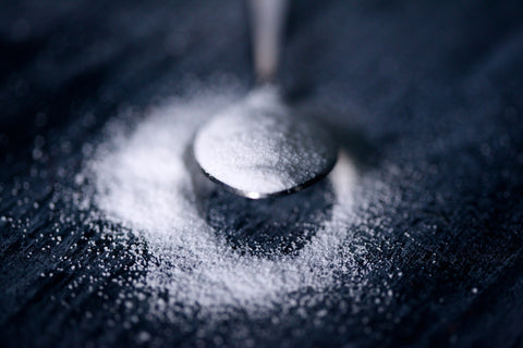 An image of a spoon full of some kind of sweeteners, poised above more of that sweetener spilled out on either side of a black surface.