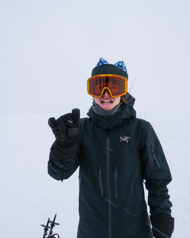 An image of a woman in ski goggles, a toque and a ski jacket waving at the camera with gloves.