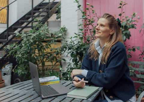 An image of a young blonde woman sitting down in front of a computer at an outside table.