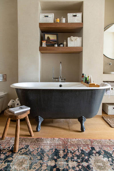 a freestanding bath tub with antique wooden stool beside it and various cosmetic products resting on a tray over the tub.