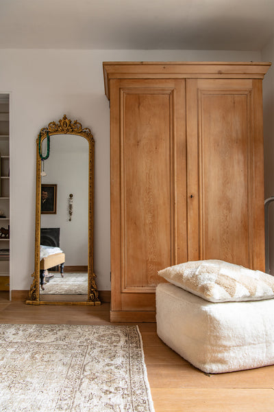 a large antique wooden armoire with a full length gilt mirror beside it. A cozy shearling pouf sits beside the cupboard.