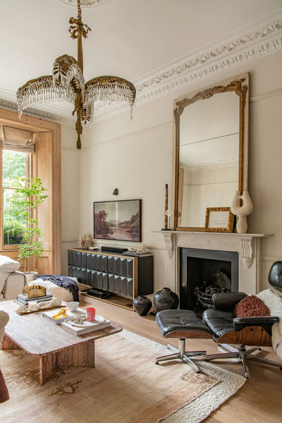 a large French antique mirror sits on top of a large marble fireplace, with a black eames chair and pink-colored Turkish rug in front.