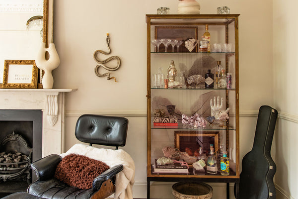 a glass cabinet with brass brackets, containing crystals and various other delicate, decorate items. A black Eames chair sits beside the cabinet, with a marble fireplace just behind it. 