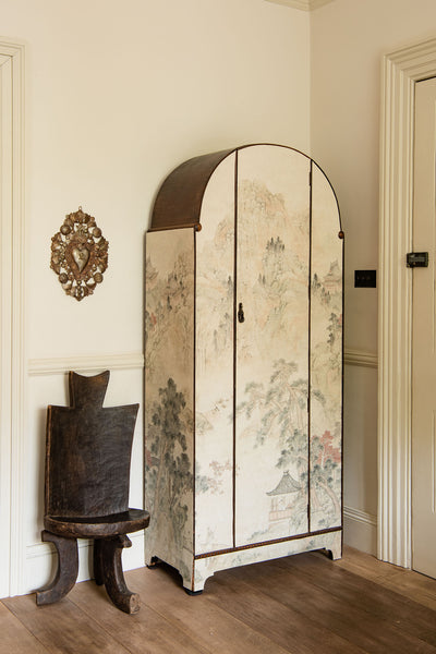 an antique wardrobe, pasted with wallpaper which depicts a washed landscape scene. Beside it sits an antique African chair, with a piece of brass religious iconography above.