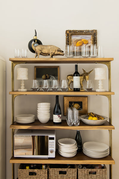 a set of open wood shelves with brass brackets, containing various glasses, artworks, crockery and wine bottles.