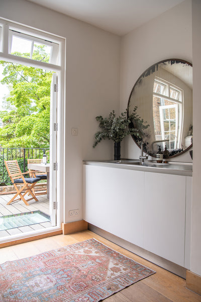 a large concave mirror hangs above a kitchen sink, with an antique rug beside it. There are two French doors leading out onto a wooden patio.