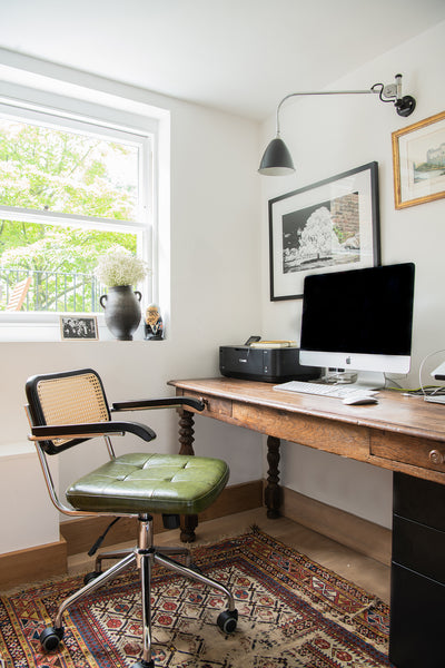 a cozy home office with an antique rug, green leather office chair and vintage wooden table, with a computer and other office items lying on top. 
