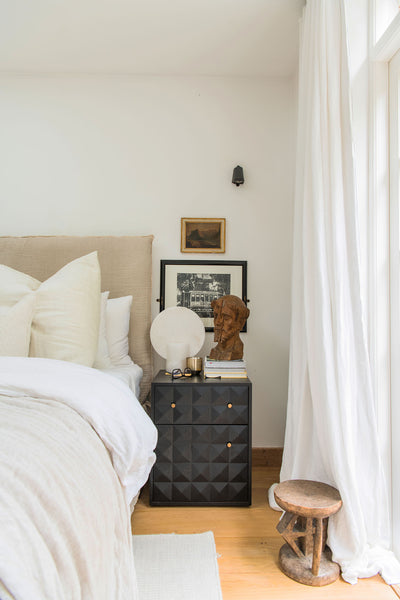 a black bedside table with various antique trinkets on top. A long white linen curtain hangs to the right, and the corner of the bed is visible to the left.