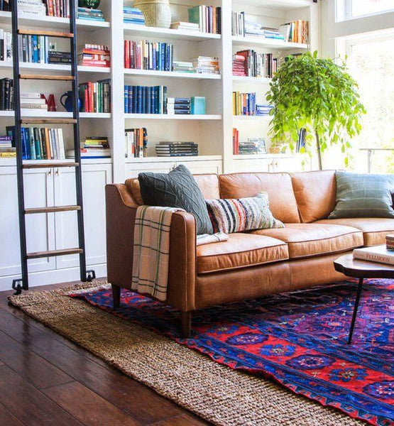 a bright red and blue antique rug layered over a brown jute. A large brown leather sofa sits above it and a library ladder is visible at the back