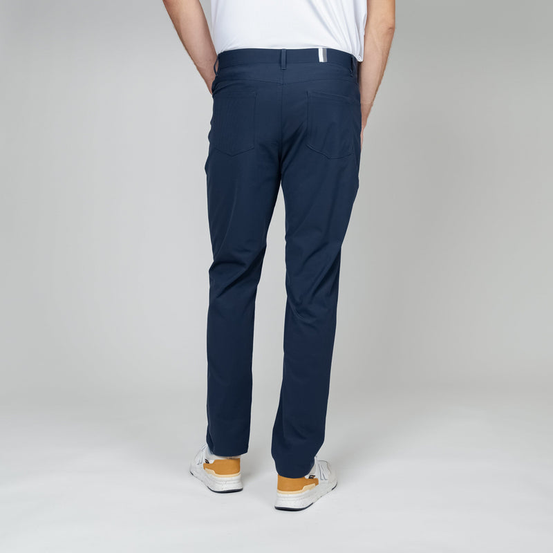 A person faces away from the camera, wearing RHOBACK's Delta Performance Pant | Solid - Fleet Navy, made from moisture-wicking, wrinkle-free fabric, paired with a white shirt and white sneakers with yellow accents against a light gray background.