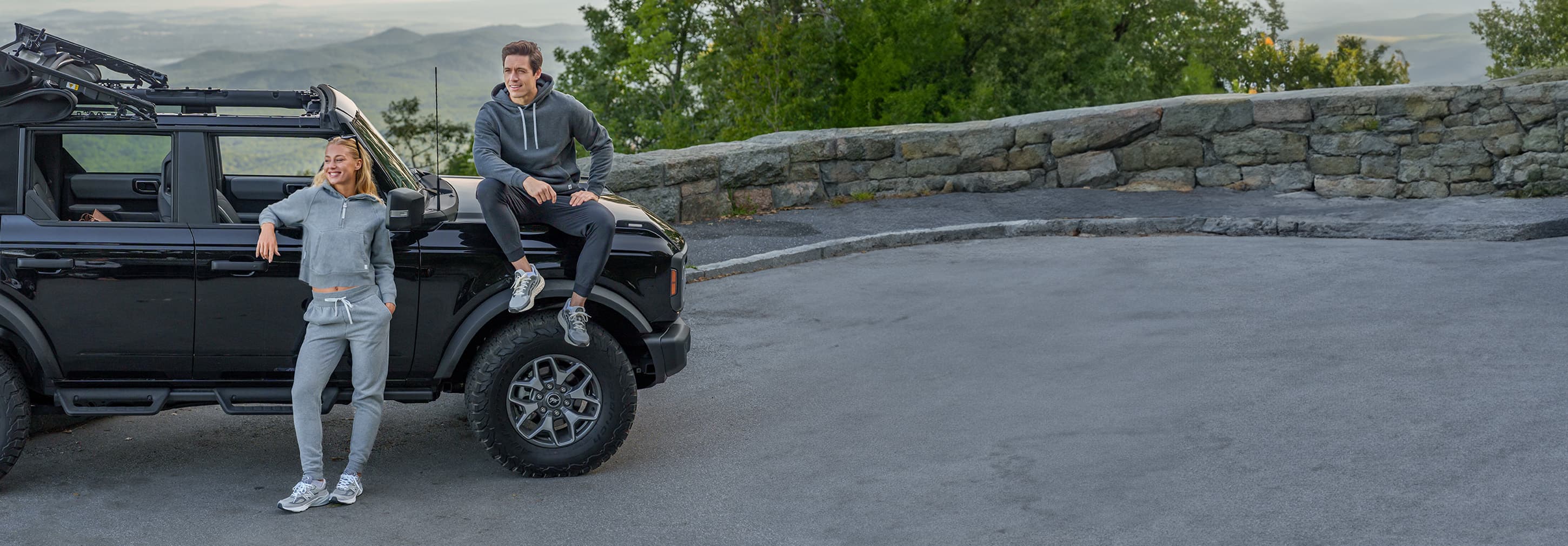 A black SUV with two people wearing Rhoback apparel sitting on its back, engaged in conversation in a scenic setting.