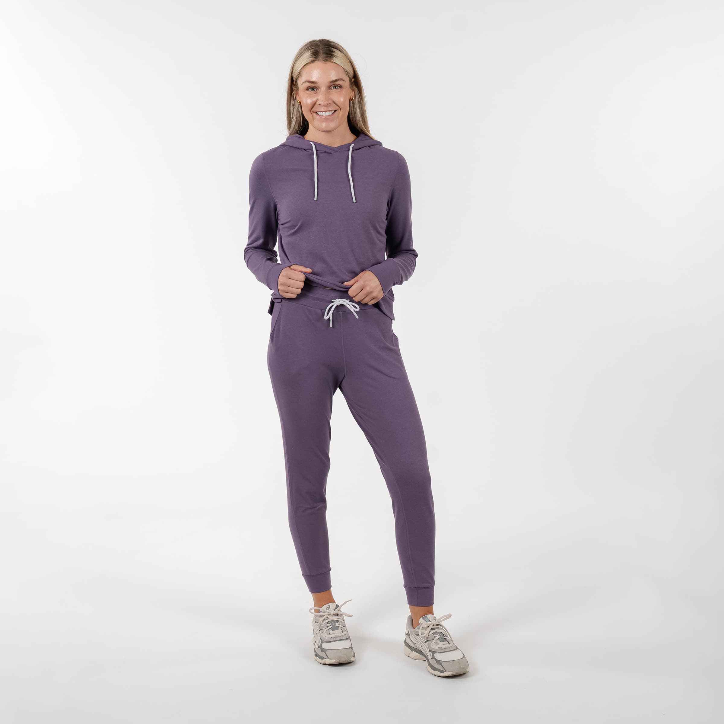 Standing in front of a plain white background, a person smiles while wearing Rhoback's Heather - Mauve Purple Jogger Set with a drawstring. They have on light-colored sneakers, and their hands rest relaxed by their sides.