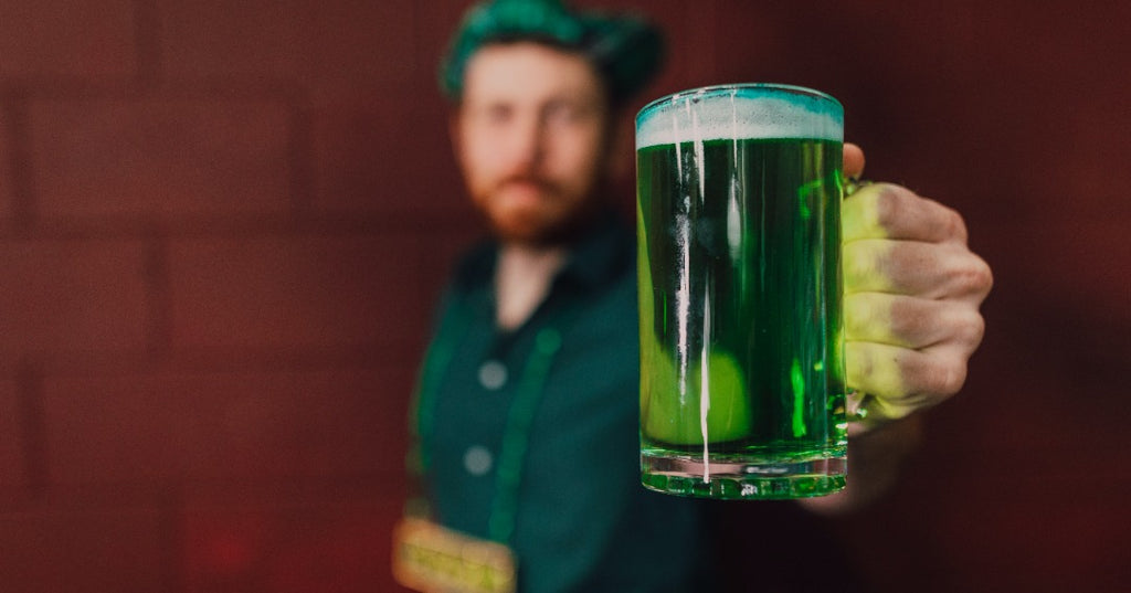 A man hold up a pint of green beer against a brick wall. the man is out of focus and dressed in St.Patrick's Day attire.