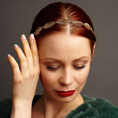 woman wearing gold leaf headband with red hair and hand by face looking down