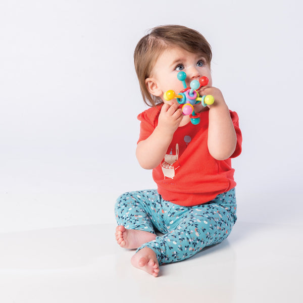 Baby girl holding and chewing on an Atom Teether toy while seated. Follow the embedded link to visit the Atom Teether toy's product page.