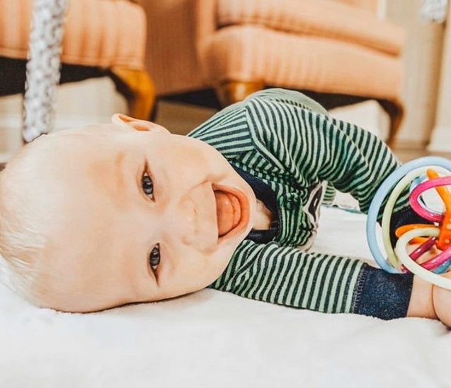 White baby boy rolling sideways on the floor with a mouth-open smile, holding a Winkel teether toy in his hands as he lays on a playmat. Photo credit to @babyledeverything on instagram.