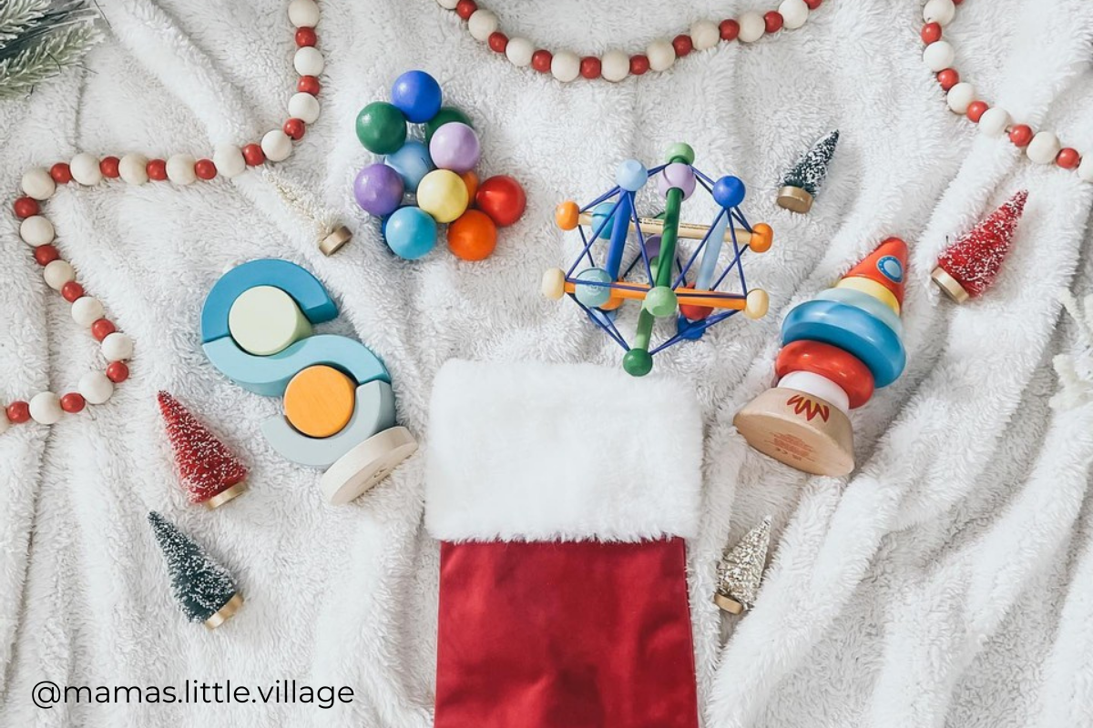 Flat lay spread of wood infant and toddler toys near stocking and holiday decor. Includes photo credit @mamas.little.village.