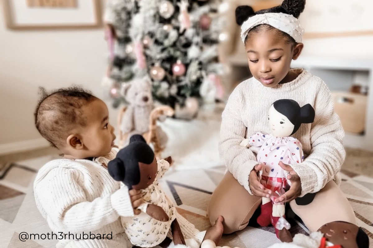 Sisters seated in front of Christmas tree with soft dolls in hand and toys all around. Photo credit to @moth3rhubbard.