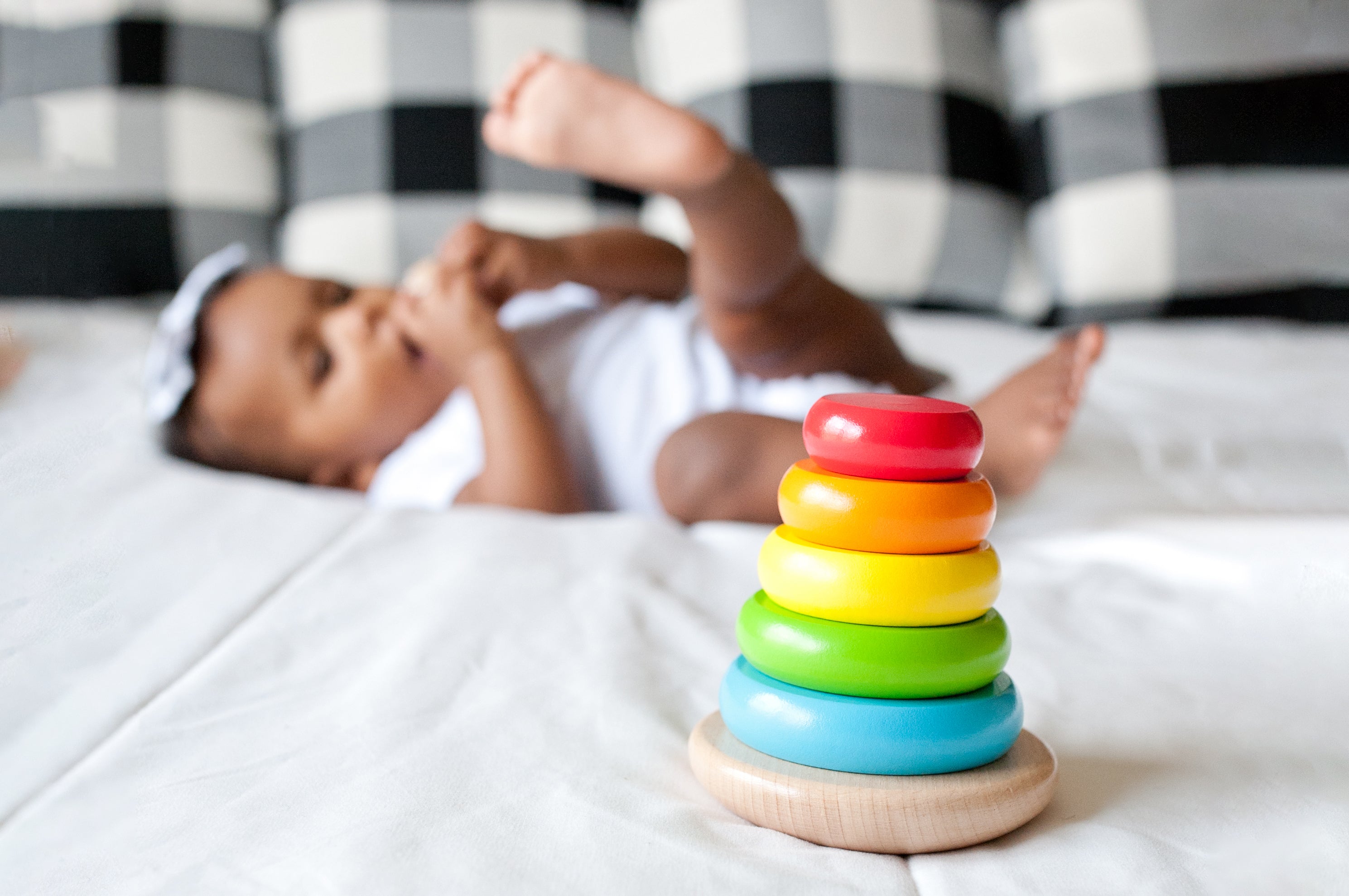 Multi-colored wood stacking toy featured prominently with background blurred. Baby girl in background holding and chewing on one of the stacking toy pieces. Photo credit to @arekeworkuphotography on Instagram.