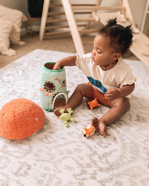 Little girl playing with fill and spill toy on rug with play gym in the background. Photo credit to Instagram user @5littlebirds_.