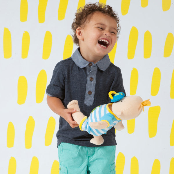Toddler-age boy holding soft baby doll while laughing. Boy is standing in front of a yellow-dash patterned wall. 