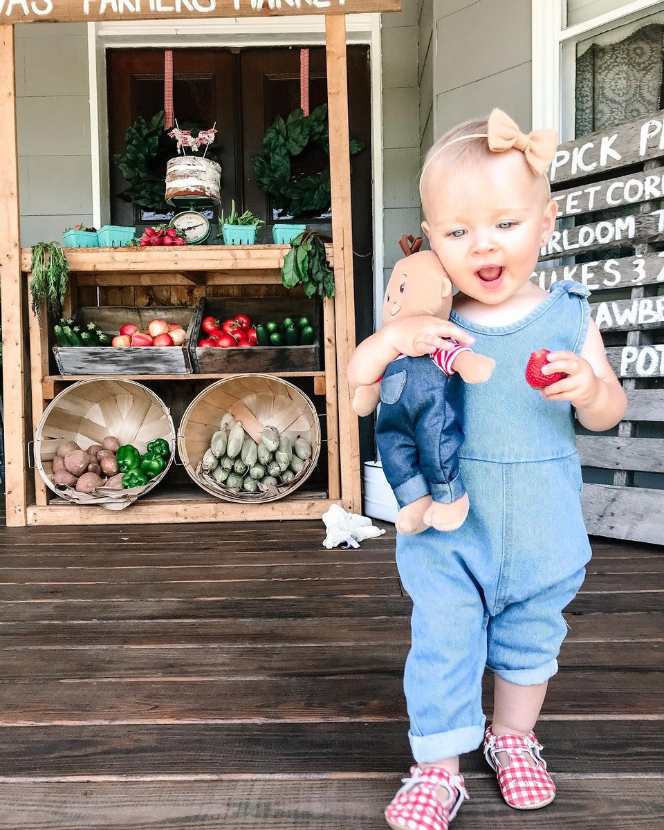 Toddler-age girl on porch with Wee Baby Stella farmer doll in hand and farmer's market stand behind her. 