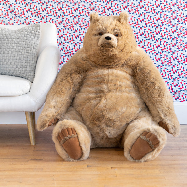 Extra large stuffed animal bear in room scene, seated on a wood floor next to a chair in front of a patterned wall.
