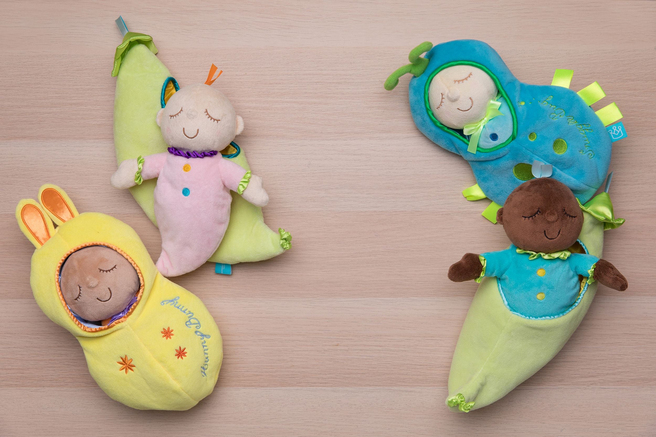 Four snuggle pod dolls laid on a wood floor backdrop. Soft dolls each have their own fabric pods that they tuck inside.