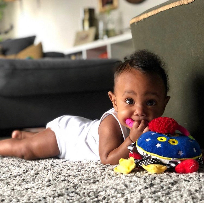 Black baby girl laying on her tummy looking straight forward with one of the arms of the Baby Whoozit toy in her mouth. Photo credit to @ocean_akin on Instagram.
