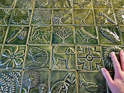 4x4 handmade tiles in leaf green glaze depicting plants and animals with the artist's hand in the frame for scale