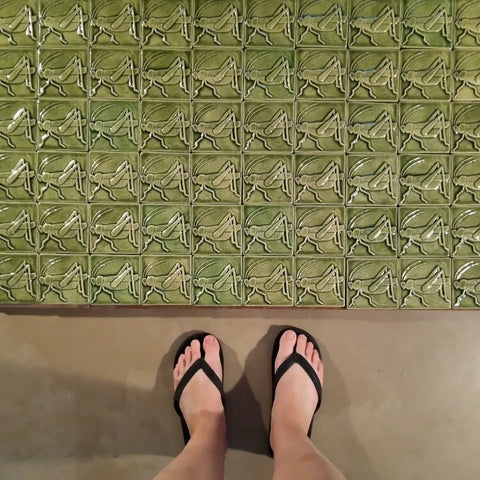 a large number of 4x4 inch, light green handmade tiles depicting grasshoppers or crickets with the artist's feet in the frame for size reference