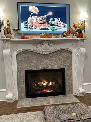 An light colored, ornate fireplace featuring carved wood and handmade tiles with thanksgiving decorations on the mantle and a fire burning in the firebox