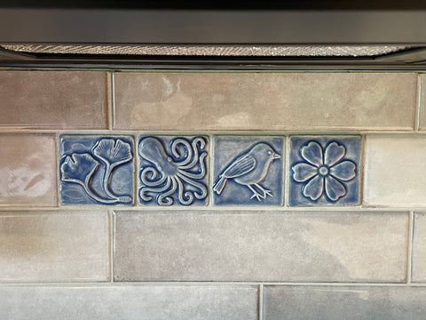 four blue handmade tiles featuring ginkgo leaves, an octopus, a chickadee and a blossom installed over a stove in a field of commercial tiles